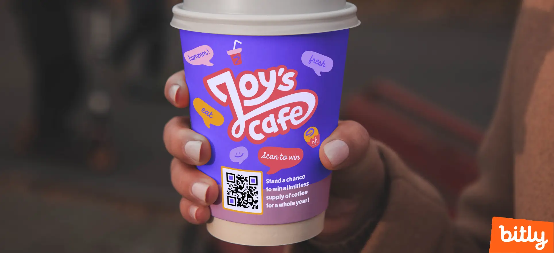 A person with painted nails holds Joy’s Cafe’s purple-shaded takeaway cup that has a QR Code on it.